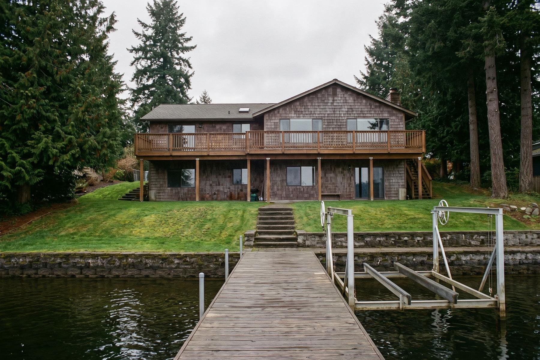 Back of a two-story cedar-shingled lake house seen from the end of its dock, with a sloping lawn, a low stone bulkhead, an empty boat lift and tall firs