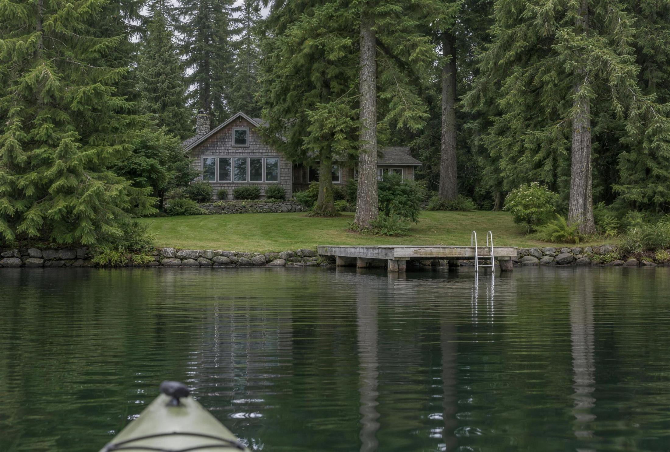 The water side of a cedar-shingled lake house seen from a kayak on an overcast morning, set back in tall firs above a sloped lawn, a low stone bulkhead, and a short wooden dock with a swim ladder